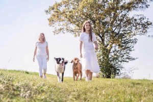Foto von Mutter und Tochter die auf einer sommerlichen Wiese mit Hunden spazieren gehen in weissen Kleidern
