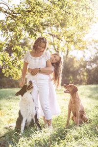 Foto von Mutter und Tochter auf einer Wiese in der Sonne mit zwei Hunden